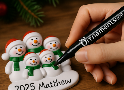 Close-up of a hand using a black ‘Ornament Shop’ marker pen to personalize a 2025 family of five snowmen Christmas ornament on a wooden surface, with festive greenery and red baubles in the background