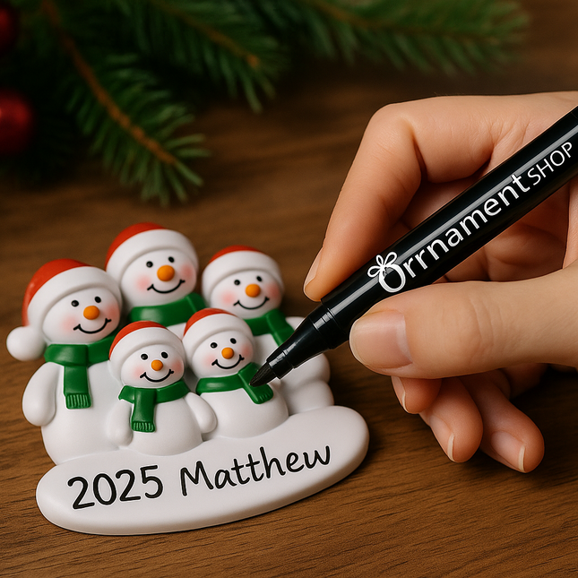 Close-up of a hand using a black ‘Ornament Shop’ marker pen to personalize a 2025 family of five snowmen Christmas ornament on a wooden surface, with festive greenery and red baubles in the background